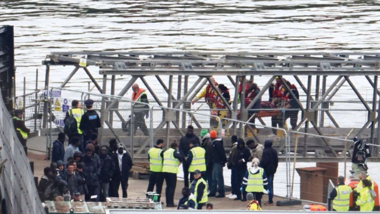 People, believed to be migrants, including one being carried on a stretcher, wait on the quayside to ...