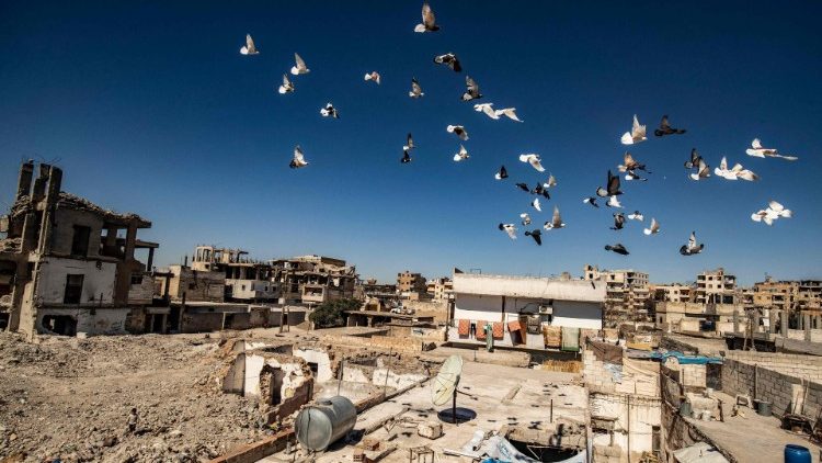 A flock of pigeons fly against a backdrop of devastation caused by ten years of conflict in Syria