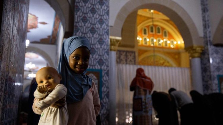 Pětileté děvčátko pózuje pro fotografii s panenkou v ruce, zatímco se muslimští věřící modlí během prvního dne měsíce ramadánu v mešitě Nizamiye v jihoafrickém Midrandu