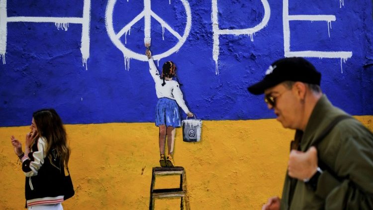 People walk past a mural with the colours of the Ukrainian flag and a sign now known as the "peace sign".