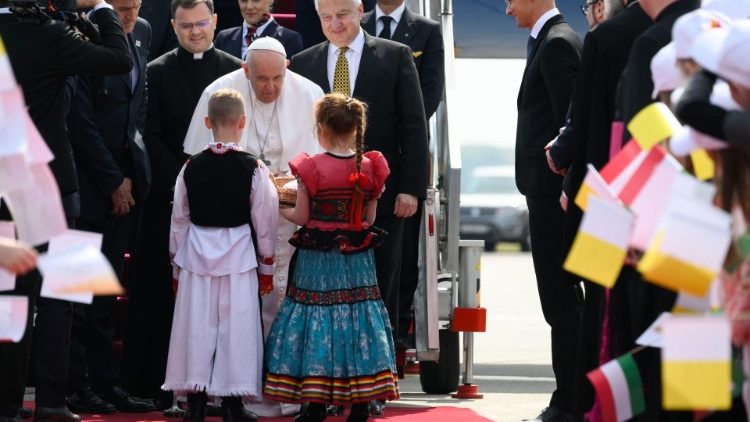 O Papa sendo acolhido no Aeroporto de Budapeste por um casal de crianças vestindo trajes típicos locais