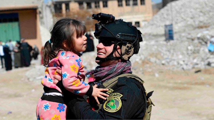 Mosul. Entre los escombros una niña mira a un soldado, con equipo de guerra, que la acaricia y la levanta, ella sonríe 