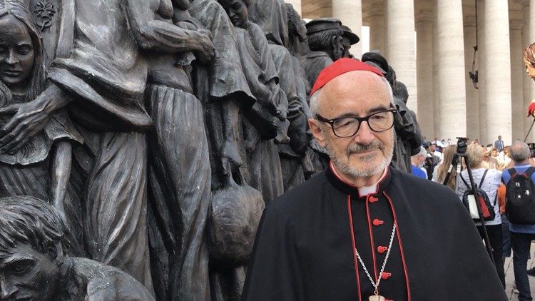 Cardinal Michael Czerny poses for a photo with a monument to refugees in St. Peter's Square