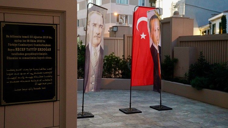 In front of the new church, banner with Mustafa Kemal Atatürk, the founder of the republic (left) and President Recep Tayyip Erdogan (right). An inscription can also be seen:This church, foundation stone laid on 03.08.2019, opening on 08.10.2023, was made possible by President Recep Tayyip Erdogan. May it be to the well-being of our country and religious community. 